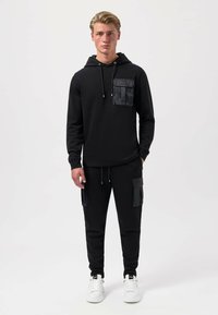 Felpa nera con tasca sul petto lucida e stringhe, abbinata a pantaloni jogger neri con tasche laterali. Scarpe da ginnastica bianche completano il look.