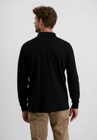 Mann mit braunen Haaren, der ein schwarzes langärmliges Poloshirt und beige Cordhosen trägt, steht mit dem Rücken zu einem schlichten Hintergrund.