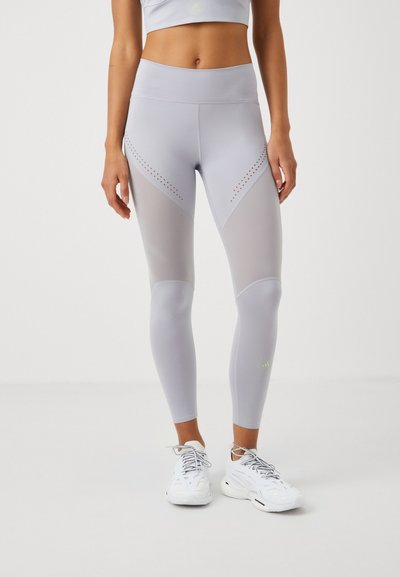 Lichtgrijze leggings met een hoge tailleband, voorzien van meshpanelen en geperforeerde ontwerpen aan de zijkanten, gecombineerd met witte sportschoenen.