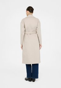 Cappotto di lana beige con vita cintrata, maniche lunghe e colletto a punte. Indossato sopra pantaloni di denim blu e scarpe nere, visto da dietro.