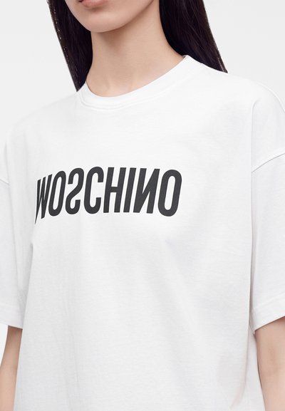 Άτομο που φοράει λευκό T-shirt με μαύρο ανάποδο και στιλιζαρισμένο κείμενο στο στήθος σε μονόχρωμο φόντο.