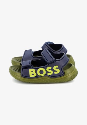 Sandales bleu marine avec semelles en caoutchouc vert, dotées de deux bandes Velcro ajustables et d'un logo "BOSS" jaune bien visible sur le côté.
