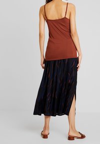 Kvinde iført en rustfarvet ærmeløs top, nederdel med mønster i marineblå og brun til midi-længde med sideslids, samt brune lavhælede slip-on sko står op.