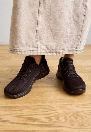 Osoba w czarnych wsuwanych tenisówkach Skechers i beżowej spódnicy, stojąca na drewnianej podłodze.