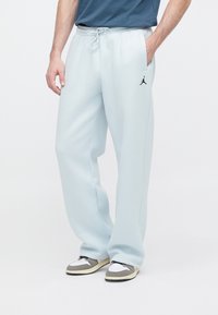 Άνδρας που φοράει ανοιχτό γαλάζιο παντελόνι φόρμας με μαύρο λογότυπο Jumpman στο αριστερό μηρό, γκρι και λευκά αθλητικά παπούτσια, και μπλε πουκάμισο, στέκεται με το ένα χέρι στην τσέπη.