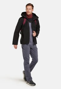 Schwarze wasserfeste Jacke mit Kapuze, graues Langarmshirt, lila Schal, graue Hose und schwarze Wanderschuhe; trägt einen Rucksack.