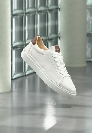 Biały skórzany sneaker z brązowymi akcentami i białymi sznurowadłami unoszący się nad błyszczącą betonową podłogą z oknami z bloków szklanych w tle.