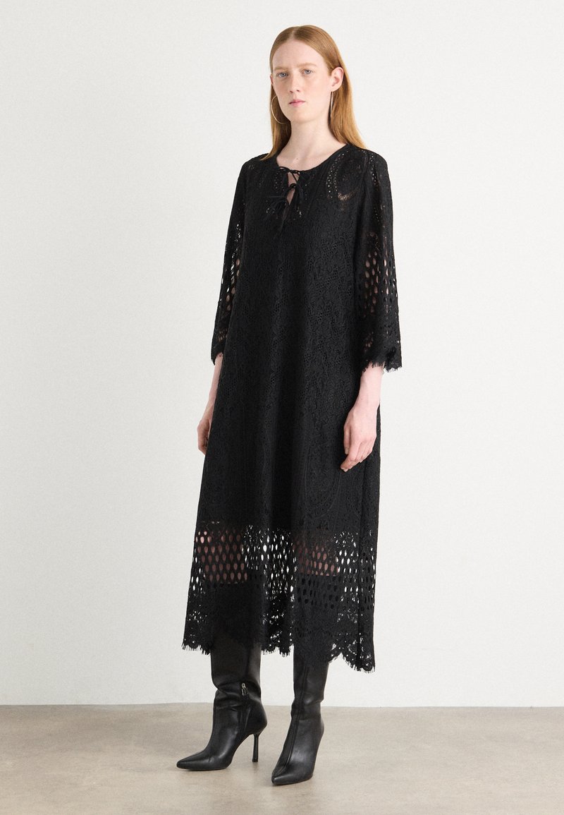 Black lace dress with three-quarter sleeves, decorative tie neckline, and an openwork hem. Worn with black heeled boots.