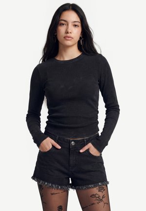 Nuori nainen, jolla on pitkät tummat hiukset, musta pitkähihainen paita, mustat farkkushortsit repsottavilla reunoilla ja läpinäkyvät sukkahousut, joissa on kukka-aiheisia tatuointeja.