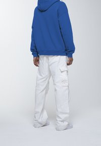 Felpa blu con cappuccio e vestibilità comoda, abbinata a pantaloni cargo bianchi con tasche. Il modello si trova di fronte a uno sfondo neutro.