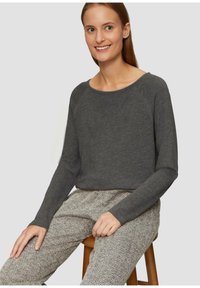 Grijze langemouwen top met een wijd uitgesneden halslijn, gecombineerd met een broek met visgraatpatroon. Model zittend op een houten kruk tegen een neutrale achtergrond.