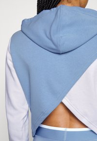 Haut à capuche court bleu et blanc avec une découpe à l'arrière, présentant une texture douce, une capuche et un design en blocs de couleur diagonaux à l'arrière.