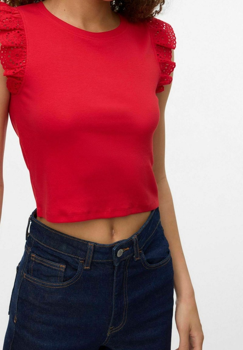 Femme portant un crop top rouge avec des manches à œillets volantées et un jean bleu foncé taille haute, sur un fond clair uni.