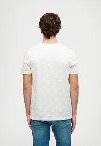 Bărbat cu părul creț, îmbrăcat cu un tricou alb cu model și mâneci scurte, și blugi albaștri, întors cu fața de la un fundal deschis.