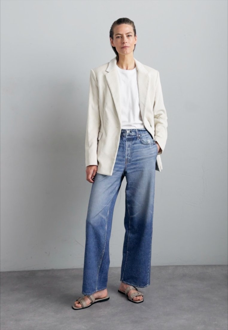 Blazer à rayures blanches sur un t-shirt blanc, associé à un jean bleu dégradé à jambes larges et des sandales plates avec des sangles métalliques. Fond léger.