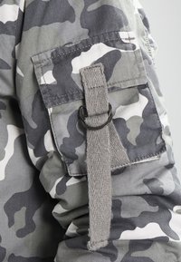 Tejido de camuflaje gris con un bolsillo de parche, detalle de correa gris y herrajes plateados. Textura suave, diseño casual y patrón variado en tonos de gris.