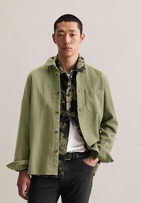 Light green button-up shirt with a textured fabric, layered over a black and green camouflage patterned shirt, paired with dark pants.