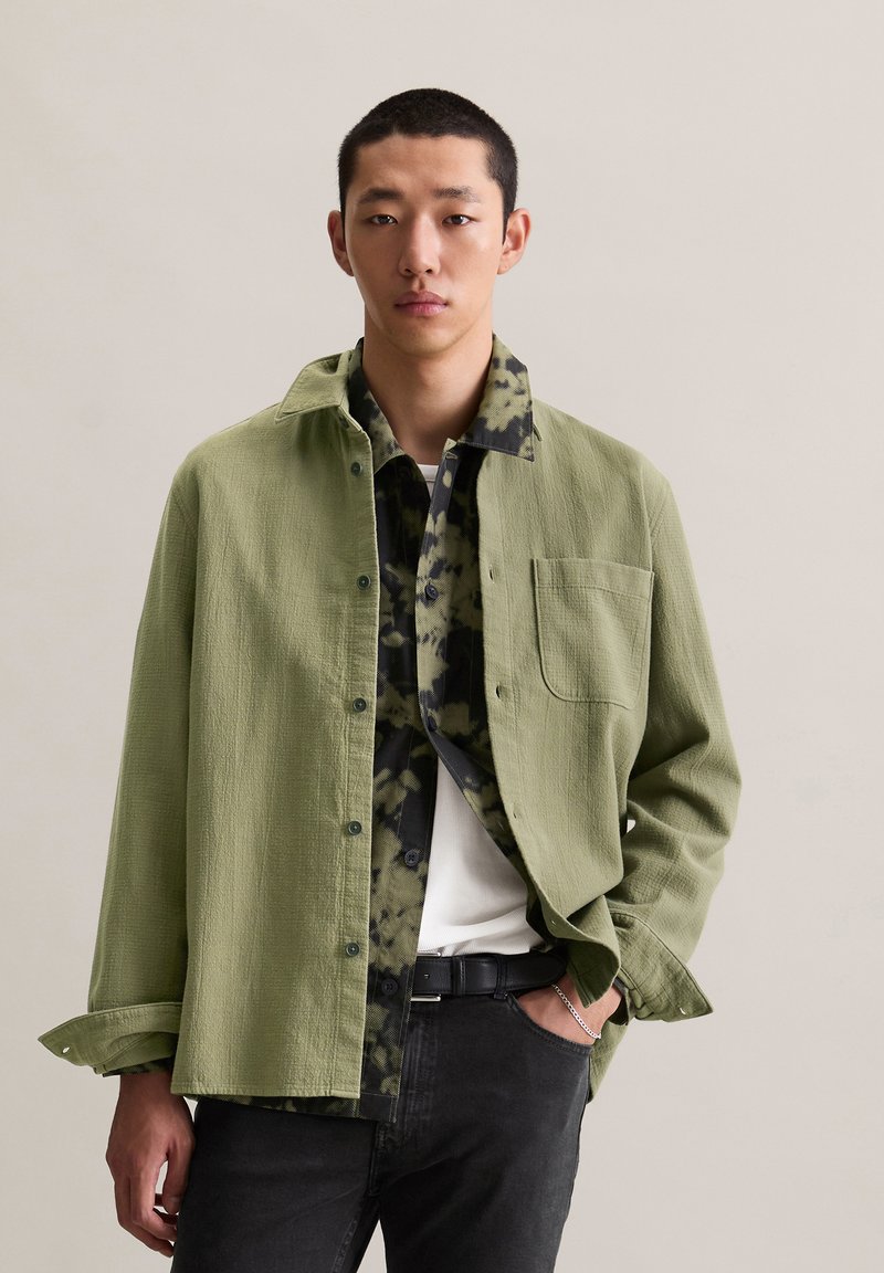 Light green button-up shirt with a textured fabric, layered over a black and green camouflage patterned shirt, paired with dark pants.