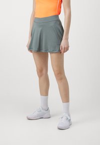 Athletic skort in muted green, made of stretch fabric. Flared design features a wide waistband, paired with white sneakers and socks.