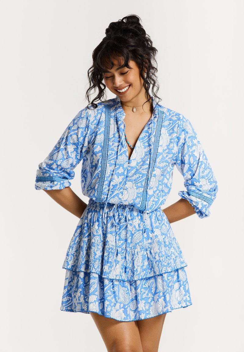Robe bleue à motifs avec une jupe à volants, ornée de dessins floraux blancs, un décolleté plongeant et des manches retroussées. Détail de tissu texturé présent.