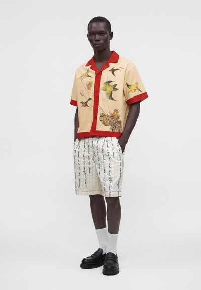 Homme portant une chemise beige et rouge à manches courtes avec un motif d’oiseaux et de paniers, un short blanc avec un texte manuscrit bleu, des chaussettes blanches et des chaussures noires.