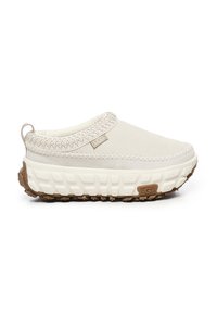 Zapato beige sin cordones con parte superior texturizada, un borde trenzado y una suela de goma gruesa y estriada. Presenta una etiqueta con el logotipo de la marca en el costado.