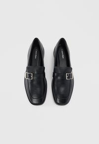 Mocassini in pelle nera con punta quadrata e fibbie argentate, marchiati Calvin Klein all'interno delle scarpe, su uno sfondo grigio chiaro.