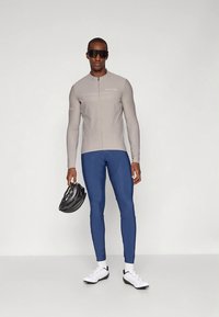 Lichtgrijze lange mouwen fietsshirt met rits, marineblauwe legging, witte sportschoenen en zwarte helm, tegen een effen achtergrond.