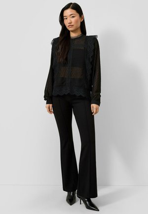 Frau mit langen dunklen Haaren trägt eine schwarze transparente Bluse mit Spitzen-Details, schwarze ausgestellte Hosen und schwarze spitze Stiefel mit hohem Absatz, steht vor einer schlichten Wand.