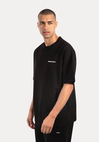 Sort bomulds T-shirt med rund hals og løstsiddende pasform, prydet med et lille hvidt "Smiledx Sport" logo på brystet.