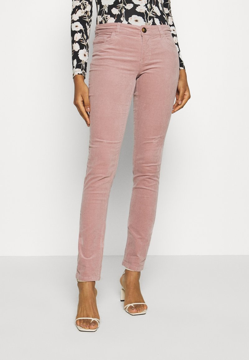 Pink corduroy pants with a fitted silhouette, featuring front pockets, a button closure, and a soft texture. Worn with a floral long-sleeve top.