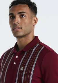 Man in een bordeauxrode polo met verticale witte, zwarte en oranje strepen en een klein pinguïnlogo op de borst.
