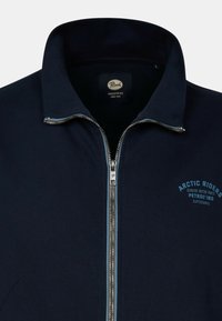 Veste zippée navy avec un col haut. Présente une fermeture éclair métallique, un texte bleu sur la poitrine gauche, et une texture côtelée autour de l'encolure.