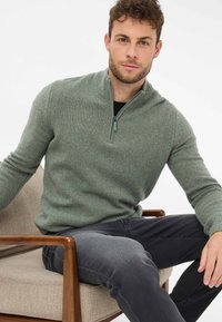 Hellgrüner Zip-Pullover mit strukturiertem Strickmuster, kombiniert mit dunklen Jeans. Model sitzt auf einem hellbeigen Stuhl.