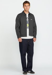 Zwarte lichtgewicht jas met een rits aan de voorkant en kraag, gedragen over een grafisch wit T-shirt. Donkerblauwe tapered jeans en zwarte sneakers.