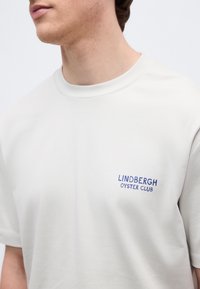 Κοντινό πλάνο ενός ατόμου που φοράει λευκό μπλουζάκι με τη φράση "LINDBERGH OYSTER CLUB" τυπωμένη με μπλε γράμματα στο στήθος.