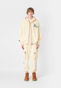 Joven vistiendo pantalones cargo crema, sudadera con cremallera a juego con un gráfico, camisa beige y botas marrones con cordones, de pie frente a un fondo blanco.