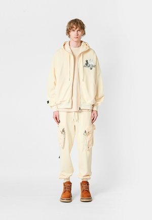 Joven vistiendo pantalones cargo crema, sudadera con cremallera a juego con un gráfico, camisa beige y botas marrones con cordones, de pie frente a un fondo blanco.