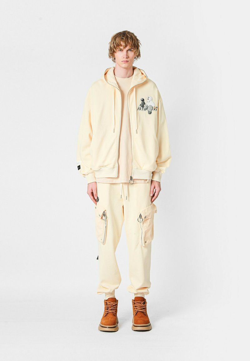 Joven vistiendo pantalones cargo crema, sudadera con cremallera a juego con un gráfico, camisa beige y botas marrones con cordones, de pie frente a un fondo blanco.