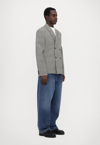 Grau karierter, doppelt geknöpfter Blazer mit breiten Revers, kombiniert mit lockeren blauen Jeans und schwarzen Schuhen, stehend vor einem weißen Hintergrund.