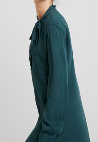 Blusa de botón en color verde azulado con cuello alto, mangas largas y un detalle de lazo en el escote. Fabricada en una tela suave y ligera.