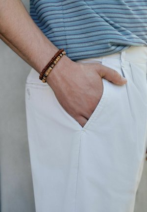 Mann trägt blau gestreiftes Hemd und weiße Hose, mit Hand in der Tasche und Perlenarmbändern am Handgelenk vor neutralem Hintergrund.