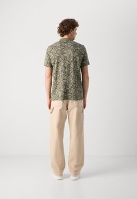 Smaragdgrünes, kurzärmliges Hemd mit Blumendruck, kombiniert mit beigen, weit geschnittenen Hosen und weißen Sneakern. Die Rückansicht zeigt die Detailnähte des Hemdes.