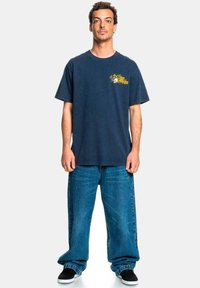Navy blue cotton t-shirt with graphic print on the left chest, paired with light blue baggy denim jeans and black slip-on shoes.
