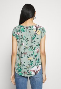 Blus med kort ärm i grönt tyg med blommönster, featuring olika flerfärgade blommor, rund halsringning och en dragkedja bak.