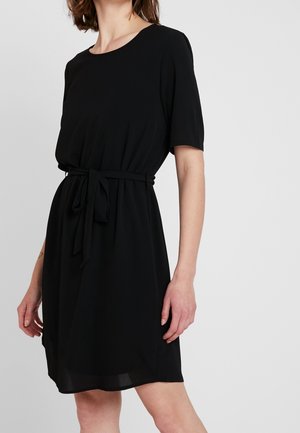 Femme portant une robe noire au genou avec manches courtes et ceinture nouée à la taille sur un fond uni.