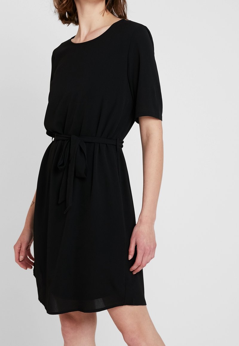 Femme portant une robe noire au genou avec manches courtes et ceinture nouée à la taille sur un fond uni.