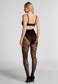 Frau trägt schwarze hoch taillierte, gemusterte Strumpfhosen, schwarzen BH, große Creolen und schwarze High Heels, mit dem Rücken zur Kamera stehend.