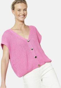 Rozā adīta jakas ar īsām piedurknēm, V veida kakla, un četriem lieliem sudraba pogām gar priekšpusi. Valkāta pār baltām biksēm.