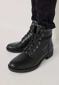 Botas de tobillo de cuero negro con cordones, ojales de metal y detalles de tachuelas en el collar. Tacón bajo tipo bloque y textura suave.